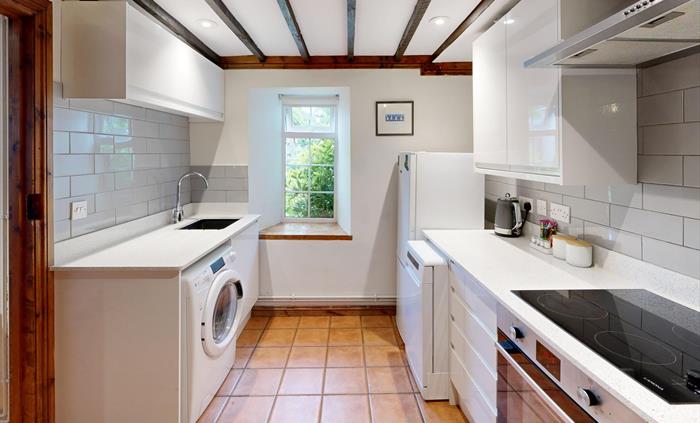 A fully-equipped kitchen with white cabinets, grey tile backsplash, terracotta floor, and a window overlooking greenery.